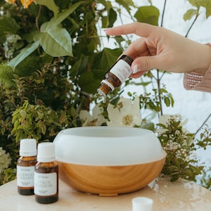 May include: A round, white, and light wood-grain essential oil diffuser sits alongside two small amber glass bottles labeled Lavender Dreams and Citrus Blossom. A third amber bottle labeled Highland Rose is held above the diffuser.