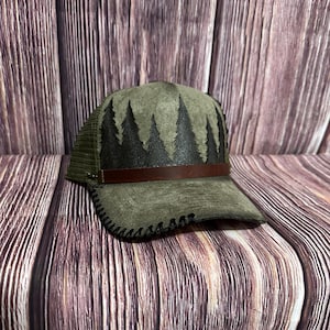 Può includere: Cappellino da camionista verde oliva con un design a forma di foresta. Il cappello presenta una fascia in pelle marrone scuro e un bordo cucito nero. Il design della foresta è realizzato in pelle marrone scuro. Il cappello ha un retro in rete.