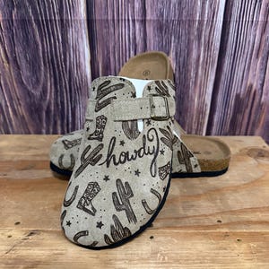 Puede incluir: Zuecos de ante color canela con temática western. Los zuecos tienen una correa con hebilla y están decorados con cactus, botas de vaquero, herraduras y la palabra "howdy". Las suelas son negras con una plantilla de corcho.