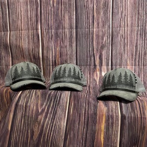 May include: Three grey trucker caps with a forest design on the front. The caps have a mesh back and a curved peak. The forest design features dark green trees. The caps are displayed on a wooden surface.
