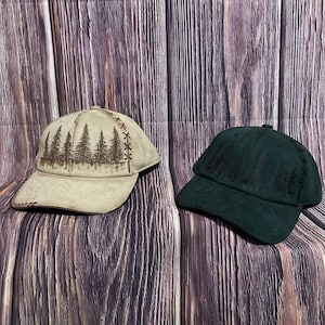 May include: Two baseball caps with a forest design. One cap is tan with a brown forest graphic, and the other is dark green with a similar forest design. Both caps have a stitched brim and a curved visor.