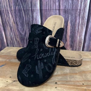 Puede incluir: Zuecos de ante negros con diseño de temática western. Los zuecos tienen cierre de hebilla y están decorados con cactus bordados, botas y la palabra "howdy". Las suelas son de color marrón claro.
