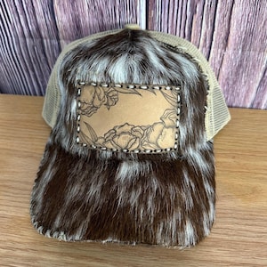 May include: A brown and white cowhide trucker hat with a brown leather patch featuring a floral design. The patch is stitched around the edges.