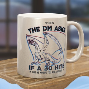 Puede incluir: Una taza de cerámica plateada metálica con una ilustración central de un dragón con efecto 3D en rojo y azul. El texto dice: WHEN THE DM ASKS IF A 30 HITS BUT HE KNOWS YOU ARE A WIZARD. La taza tiene un asa estándar y un cuerpo cilíndrico.