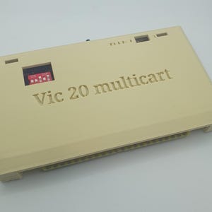 May include: A beige Vic 20 multicart with the text "Vic 20 multicart" engraved on the top. The device has a small red and black switch panel and a series of pins along the bottom edge.
