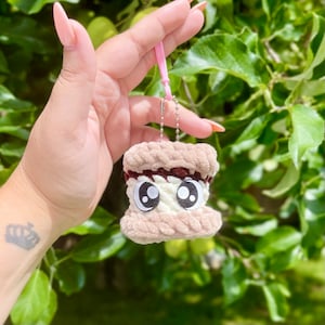 May include: A brown and white crocheted s'mores keychain with googly eyes. The keychain is attached to a pink string and a silver chain.