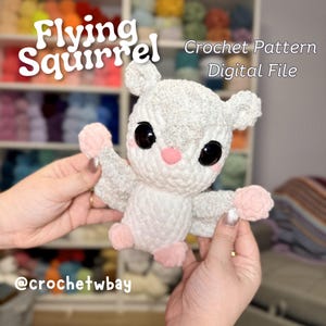 May include: A crocheted flying squirrel amigurumi toy in white with pink accents. The toy is being held by two hands. The text "Flying Squirrel Crochet Pattern Digital File" is above the toy. The text "@crochetwbay" is below the toy.
