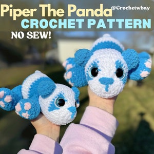 May include: Two crocheted panda plushies, one blue and white and one smaller, are held in the hands of a person wearing a pink shirt. The plushies have large black eyes and pink paw pads. The text "Piper The Panda CROCHET PATTERN NO SEW!" is at the top of the image.