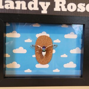 May include: A framed artwork featuring a brown, oval-shaped object with a decorative element, set against a blue sky background with white clouds. The frame is black, and the artwork is labeled "Mandy Rose".