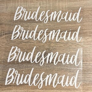 May include: Four white vinyl decals with the word "Bridesmaid" in a cursive font. The decals are arranged on a light brown wooden surface. The word is repeated four times.
