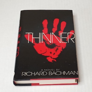May include: A hardcover book titled Thinner by Richard Bachman. The black dust jacket features a large, prominent red handprint in the center. The title is printed in thin white capital letters across the handprint, with author name below.