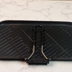 May include: A rectangular black cast iron grill pan featuring a textured surface with diagonal ridges for searing. The pan includes a built-in pouring spout on the left side and a foldable metal handle attached to the center of the bottom edge.