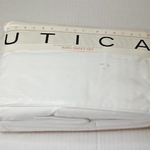 May include: A white king sheet set packaged in clear plastic. The label at the top reads LUXURY 200 PERCALE, UTICA, and KING SHEET SET in black text. The sheets are folded neatly inside the transparent wrapping, showing a solid white color.