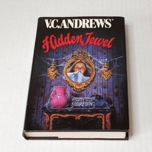 May include: A hardcover book titled Hidden Jewel by V.C. Andrews. The black cover features a dark room with a gold-framed mirror showing a person, a pink vase, a small ornate box, and a dark table, all covered in spiderwebs against purple wallpaper.