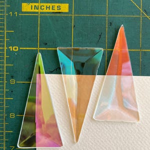 May include: Three triangular acrylic pieces with an iridescent finish that shifts between pink, orange, and teal hues. The pieces are arranged on a white surface next to a green cutting mat with a yellow grid showing measurements in inches.