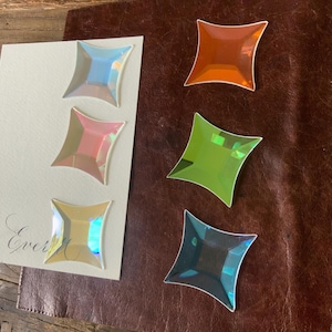 May include: Five iridescent glass squares with beveled edges. The squares are in shades of orange, pink, green, blue, and gold.