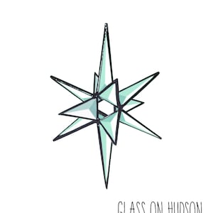 May include: A hand-made, light blue glass starburst ornament with a black outline. The ornament is labeled "GLASS ON HUDSON HAND MADE".