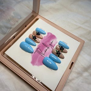 May include: A set of ten false nails in a light blue and nude colour with a black berry design. The nails are displayed in a white box with the words "Thank you" written on it.