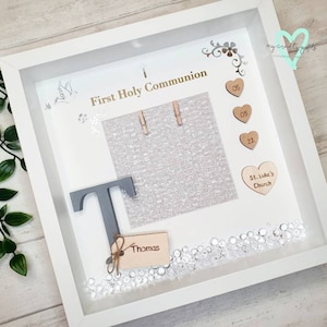 K&ouml;nnte beinhalten: Wei&szlig;e Schattenbox mit einem grauen Buchstaben T, einem Holzschild mit dem Namen Thomas und einem Fotorahmen mit dem Text "Erste Heilige Kommunion". Die Schattenbox ist mit silbernem Glitzer, Holzh Herzen mit den Zahlen 05 und 12 sowie einem Holzh Herzen mit dem Text "St. Luke's Church" verziert.