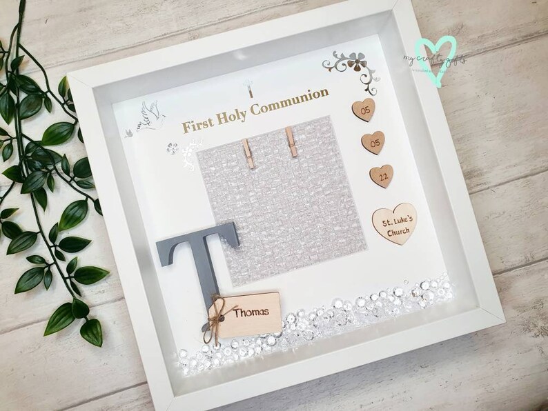 K&ouml;nnte beinhalten: Wei&szlig;e Schattenbox mit einem grauen Buchstaben T, einem Holzschild mit dem Namen Thomas und einem Fotorahmen mit dem Text "Erste Heilige Kommunion". Die Schattenbox ist mit silbernem Glitzer, Holzh Herzen mit den Zahlen 05 und 12 sowie einem Holzh Herzen mit dem Text "St. Luke's Church" verziert.