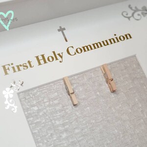 K&ouml;nnte beinhalten: Wei&szlig;er Bilderrahmen mit einer goldenen Aufschrift "Erste Heilige Kommunion". Der Rahmen hat ein silbernes Kreuz und zwei silberne Blumen in der oberen linken Ecke. Auf der rechten Seite des Rahmens befinden sich zwei Holz-W&auml;scheklammern.