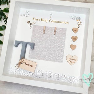 K&ouml;nnte beinhalten: Wei&szlig;er Bilderrahmen mit einem grauen Buchstaben T, einer wei&szlig;en Karte mit dem Text "Erste Heilige Kommunion" und einer wei&szlig;en Karte mit dem Text "St. Luke's Church". Der Rahmen ist mit kleinen Holzh Herzen, einer silbernen Taube und silbernen Blumen verziert.