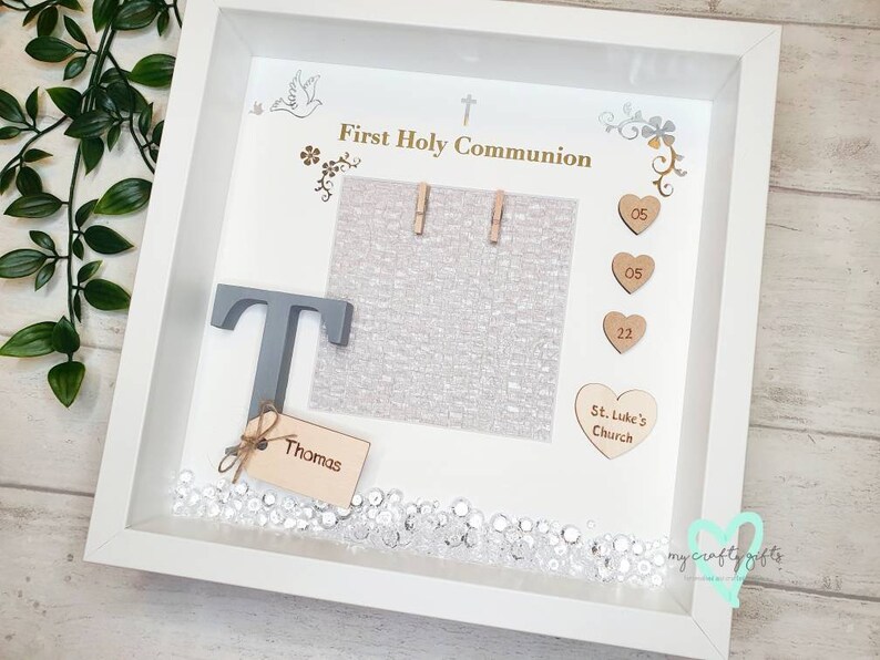 K&ouml;nnte beinhalten: Wei&szlig;er Bilderrahmen mit einem grauen Buchstaben T, einer wei&szlig;en Karte mit dem Text "Erste Heilige Kommunion" und einer wei&szlig;en Karte mit dem Text "St. Luke's Church". Der Rahmen ist mit kleinen Holzh Herzen, einer silbernen Taube und silbernen Blumen verziert.