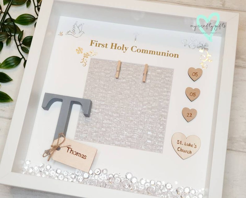 K&ouml;nnte beinhalten: Wei&szlig;e Schattenbox mit einem grauen Buchstaben T, einem Holzschild mit dem Namen Thomas und einer wei&szlig;en Karte mit dem Text "Erste Heilige Kommunion". Die Schattenbox enth&auml;lt au&szlig;erdem eine wei&szlig;e Karte mit dem Text "St. Luke's Church", drei Holzh Herzen mit den Zahlen 05, 05 und 22 sowie einen wei&szlig;en Hintergrund mit silbernem Glitzer.
