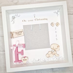 May include: White wooden frame with a photo opening, a pink letter E, a small heart with the name Emma, and a wooden figurine of a baby. The frame also includes a cross, a tag with the text "From Godparents Grigore and Cristina", and the date 2025. The text "On your Christening" is written on the frame.