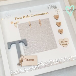 K&ouml;nnte beinhalten: Wei&szlig;e Schattenbox mit einem grauen Buchstaben T, einem Holzschild mit dem Namen Thomas und einem Fotorahmen mit dem Text "First Holy Communion". Die Schattenbox ist mit kleinen Holzh Herzen mit den Zahlen 05 und 22, einem Herz mit dem Text "St. Luke's Church" und Strasssteinen verziert.