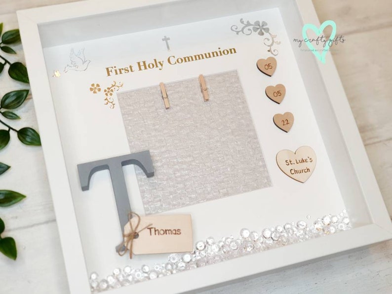 K&ouml;nnte beinhalten: Wei&szlig;e Schattenbox mit einem grauen Buchstaben T, einem Holzschild mit dem Namen Thomas und einem Fotorahmen mit dem Text "First Holy Communion". Die Schattenbox ist mit kleinen Holzh Herzen mit den Zahlen 05 und 22, einem Herz mit dem Text "St. Luke's Church" und Strasssteinen verziert.