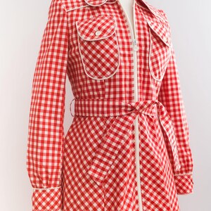 May include: A red and white gingham dress with a white zipper down the front and a white belt. The dress has two white-trimmed pockets on the chest and white trim around the collar and cuffs.