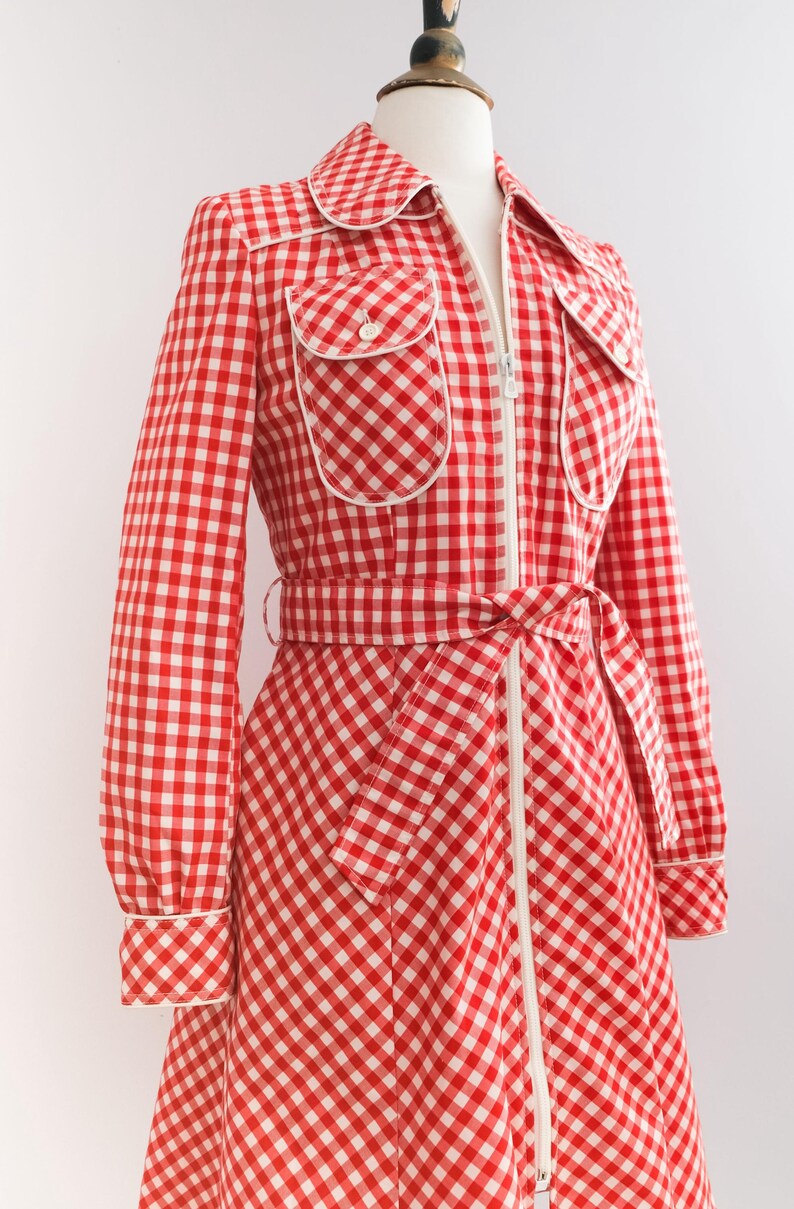 May include: A red and white gingham dress with a white zipper down the front and a white belt. The dress has two white-trimmed pockets on the chest and white trim around the collar and cuffs.