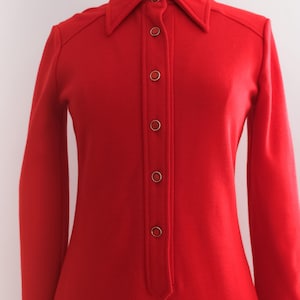 Op de afbeelding: Een felrode vintage jurk met lange mouwen en een kraag. De jurk heeft een knoopsluiting aan de voorkant met zes knopen. De jurk wordt gepresenteerd op een mannequin tegen een effen witte achtergrond. De jurk lijkt gemaakt van een zachte, geweven stof.