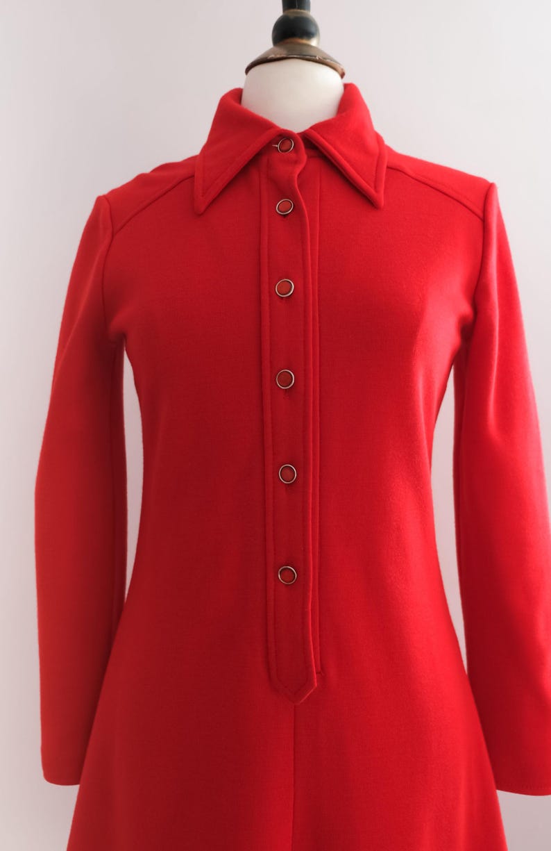 Op de afbeelding: Een felrode vintage jurk met lange mouwen en een kraag. De jurk heeft een knoopsluiting aan de voorkant met zes knopen. De jurk wordt gepresenteerd op een mannequin tegen een effen witte achtergrond. De jurk lijkt gemaakt van een zachte, geweven stof.