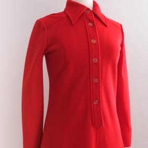 Op de afbeelding: Een vintage rode jurk met een kraag en lange mouwen. De jurk heeft een knoopsluiting aan de voorkant met goudkleurige knopen. De jurk wordt gepresenteerd op een mannequin, die het klassieke ontwerp en silhouet laat zien.