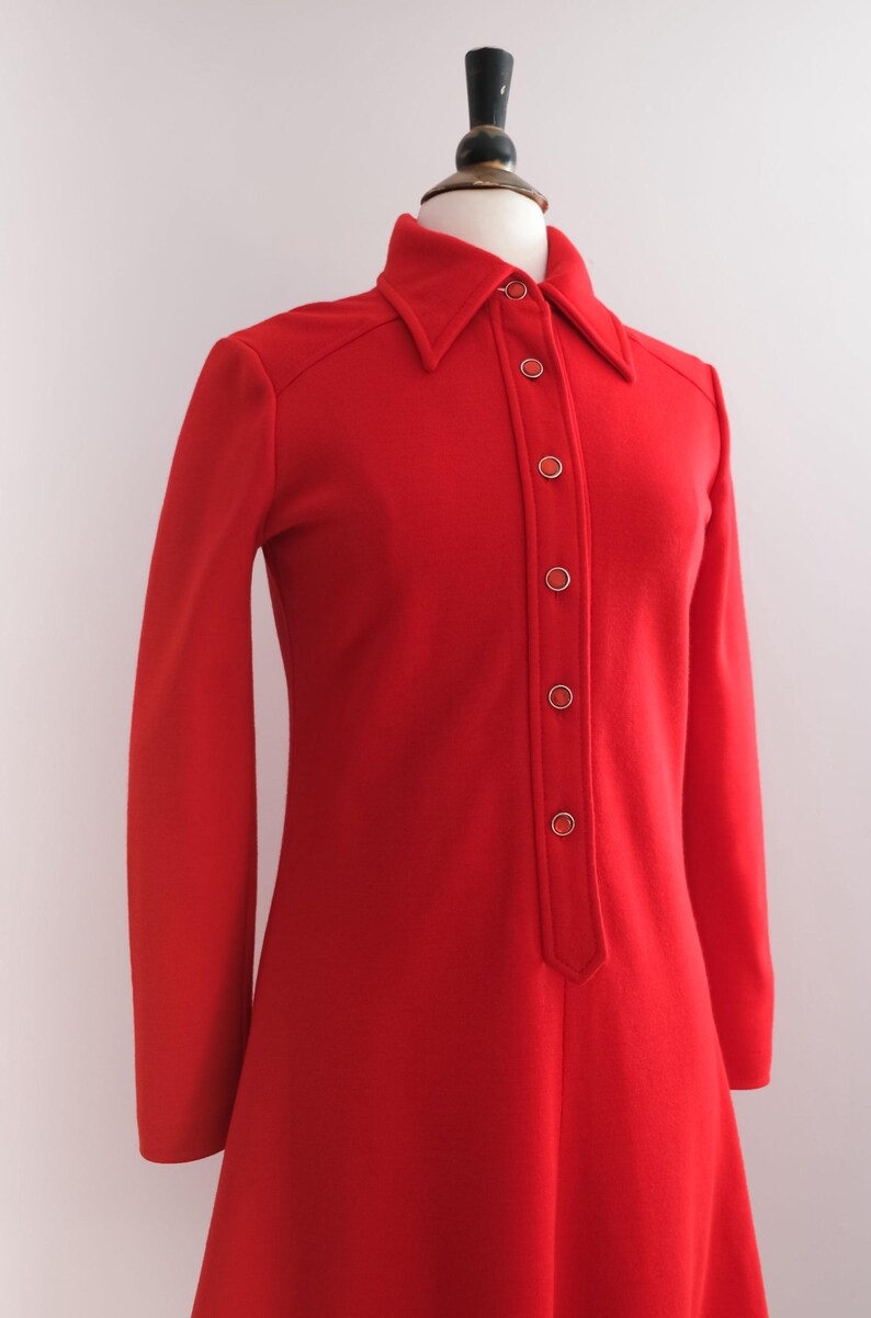 Op de afbeelding: Een vintage rode jurk met een kraag en lange mouwen. De jurk heeft een knoopsluiting aan de voorkant met goudkleurige knopen. De jurk wordt gepresenteerd op een mannequin, die het klassieke ontwerp en silhouet laat zien.