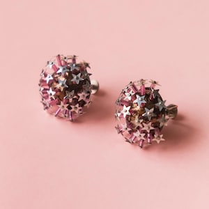 May include: A pair of vintage earrings. Each earring is a sphere with a silver star design and pink accents. The earrings have a post backing and are set against a pink background.