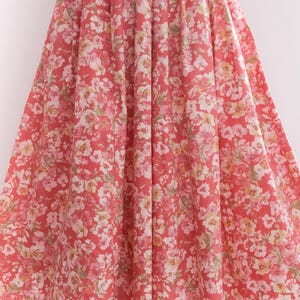 May include: A full-length, flowing dress with a vibrant floral pattern. The dress features a coral-red background adorned with clusters of white, pink, and yellow flowers. The fabric is gathered at the waist, creating a full skirt.