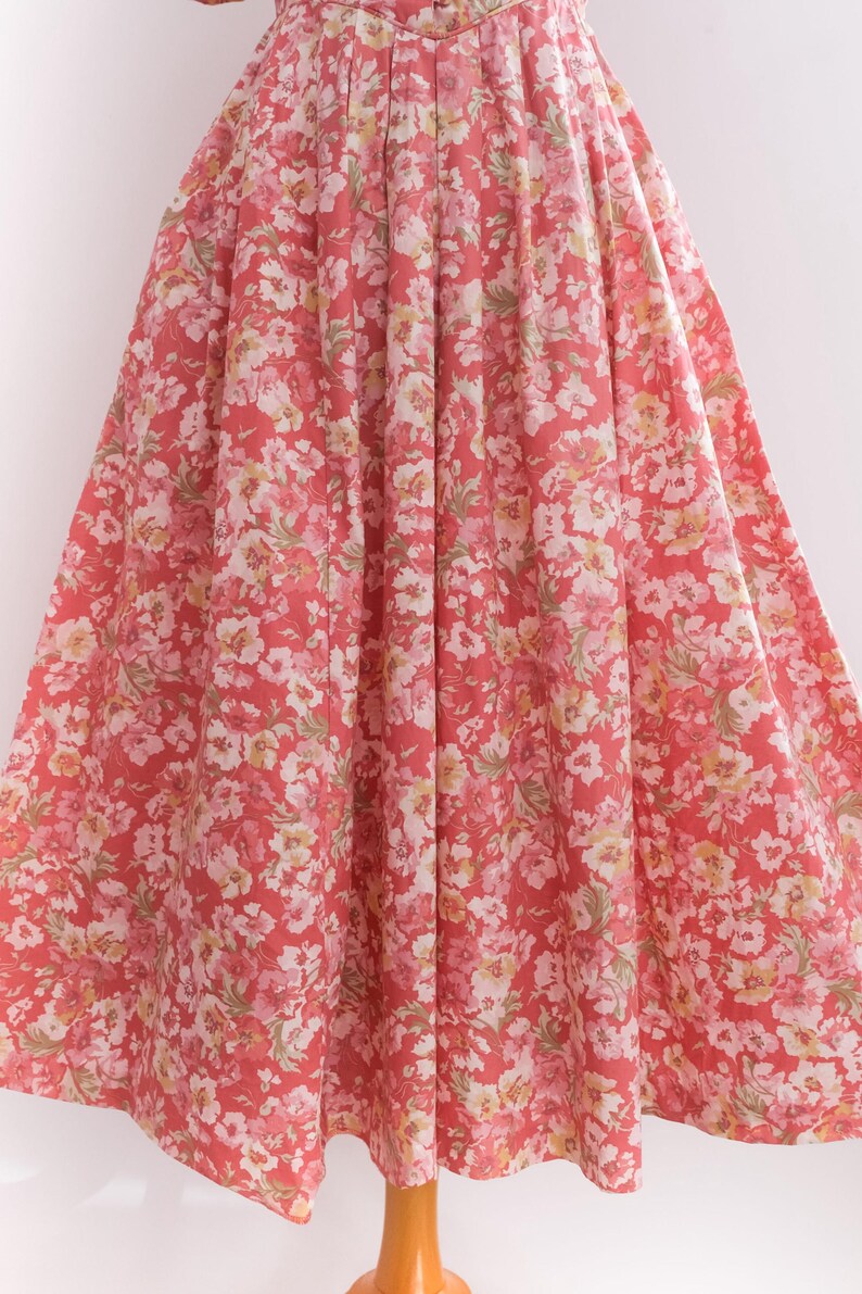 May include: A full-length, flowing dress with a vibrant floral pattern. The dress features a coral-red background adorned with clusters of white, pink, and yellow flowers. The fabric is gathered at the waist, creating a full skirt.