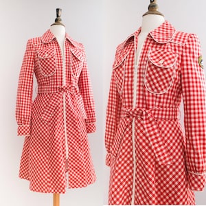 May include: A red and white gingham dress with a white zip down the front and a white belt. The dress has two white-trimmed pockets on the chest and a white trim around the collar.
