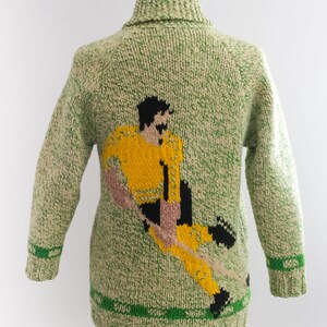 May include: A green and cream-colored knitted sweater featuring a soccer player in yellow, black, and pink. The sweater has a shawl collar and green stripes on the cuffs and hem. The design is a unique, handmade piece.