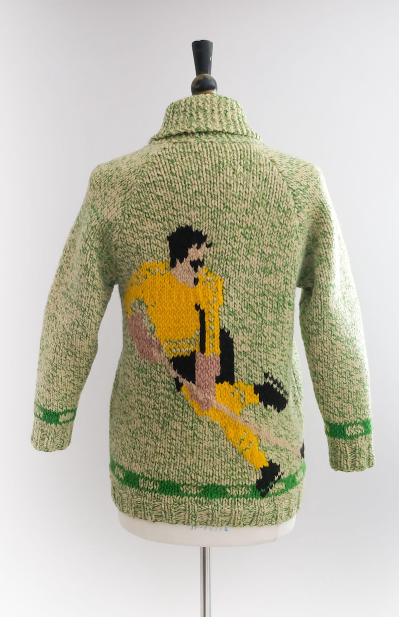 May include: A green and cream-colored knitted sweater featuring a soccer player in yellow, black, and pink. The sweater has a shawl collar and green stripes on the cuffs and hem. The design is a unique, handmade piece.
