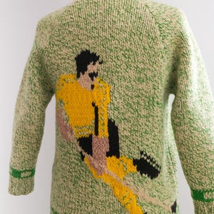May include: A green and cream-colored knitted sweater featuring a soccer player in yellow, black, and tan. The sweater has a collared neckline and green stripes on the cuffs and hem. The soccer player is in mid-kick.