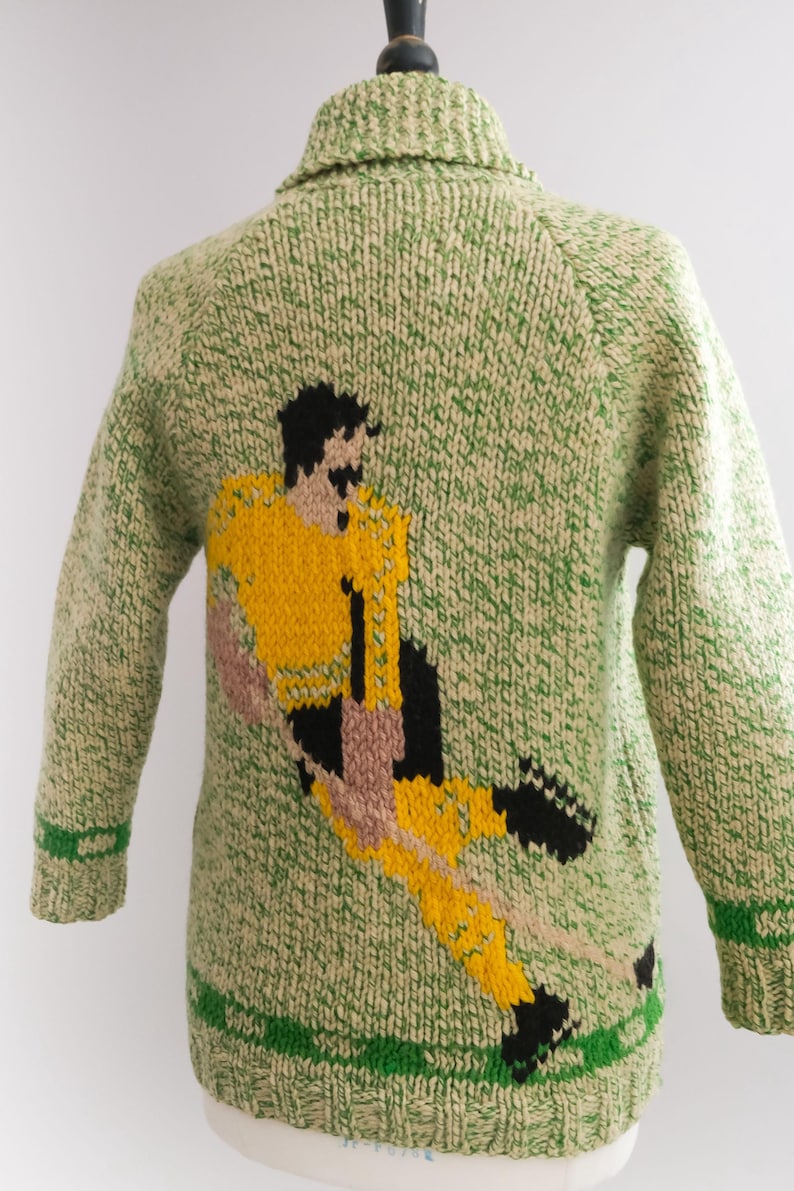 May include: A green and cream-colored knitted sweater featuring a soccer player in yellow, black, and tan. The sweater has a collared neckline and green stripes on the cuffs and hem. The soccer player is in mid-kick.