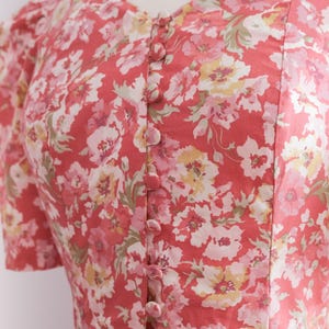 May include: A coral-colored dress with a floral pattern featuring pink, white, and yellow flowers. The dress has a V-neckline, short sleeves, and a row of matching buttons down the front. The fabric appears to be lightweight.