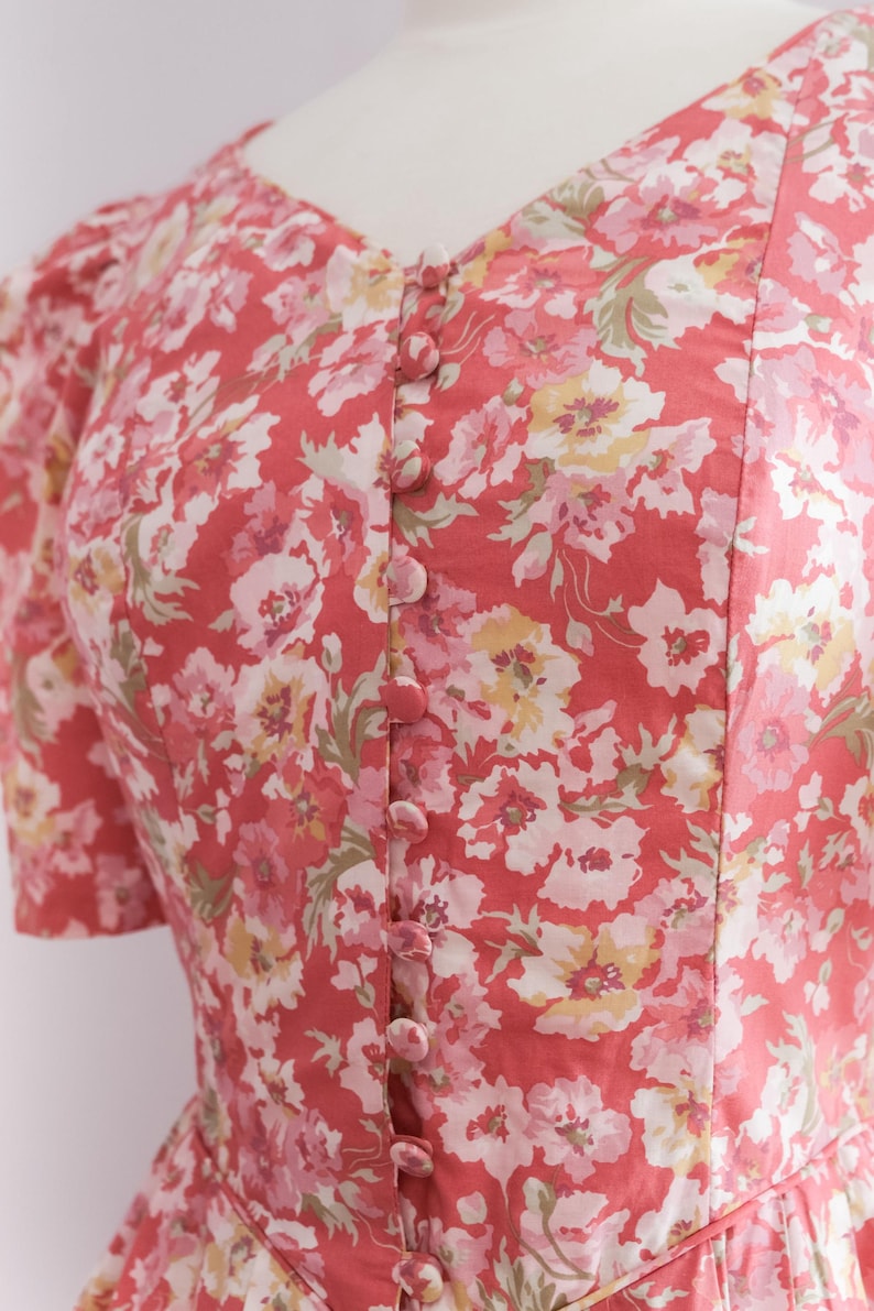 May include: A coral-colored dress with a floral pattern featuring pink, white, and yellow flowers. The dress has a V-neckline, short sleeves, and a row of matching buttons down the front. The fabric appears to be lightweight.