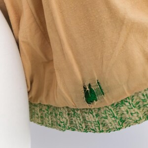 May include: Close-up of a tan fabric with a green and white knitted trim. The fabric has a tear revealing a green interior. The knitted trim is a thick, textured pattern.