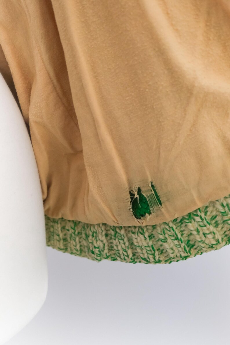 May include: Close-up of a tan fabric with a green and white knitted trim. The fabric has a tear revealing a green interior. The knitted trim is a thick, textured pattern.