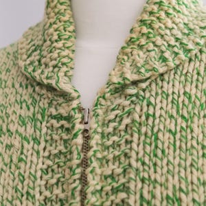 May include: Close-up of a cream-colored knitted jacket with green accents. The jacket features a high collar and a full zipper closure. The thick knit creates a textured appearance, and the green yarn is interspersed throughout the cream base.